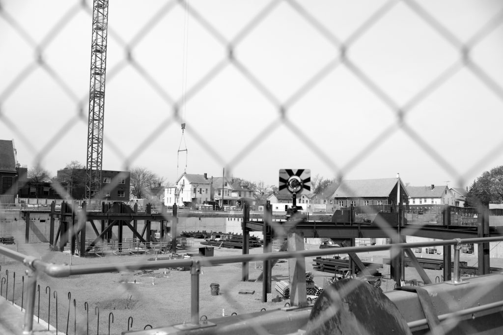Black and white photo of the construction site through a chainlink fence.