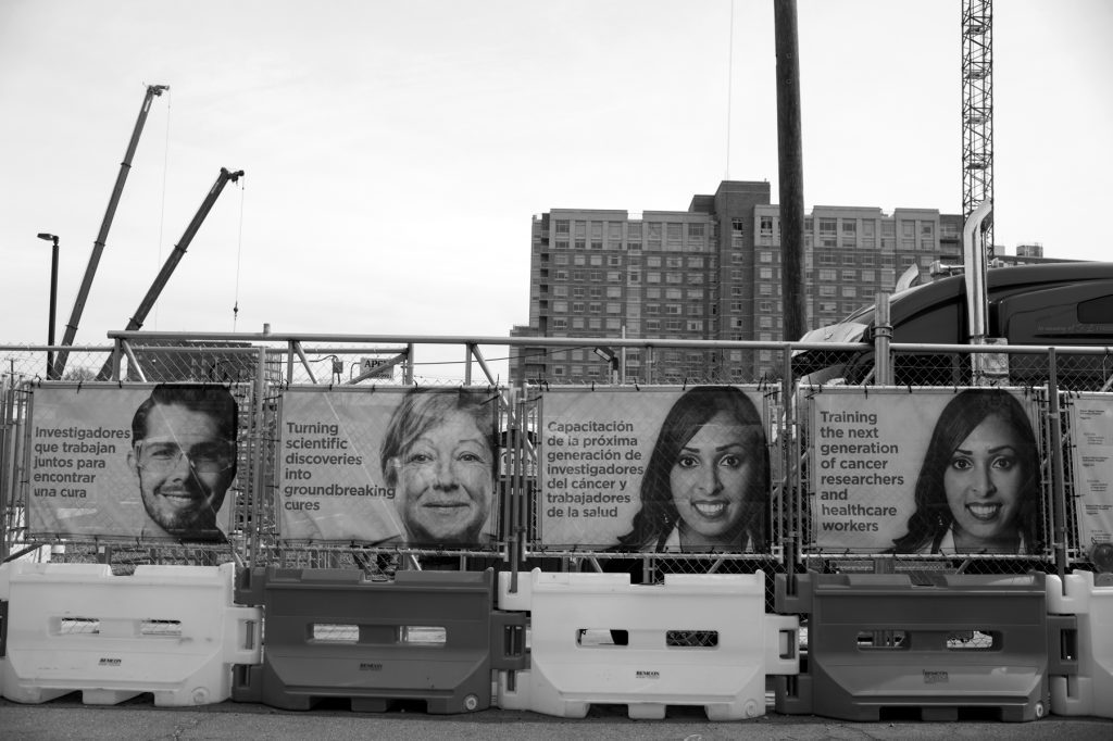 Black and white photo of ads lining the fence of the construction site, showing smiling faces and copy in English and Spanish: "Training the next generation of cancer researchers and healthcare workers" 